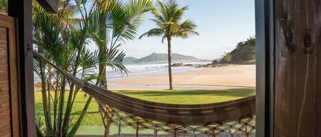 A hammock on a wooden deck overlooking a beach with palm trees and mountains in the distance.
