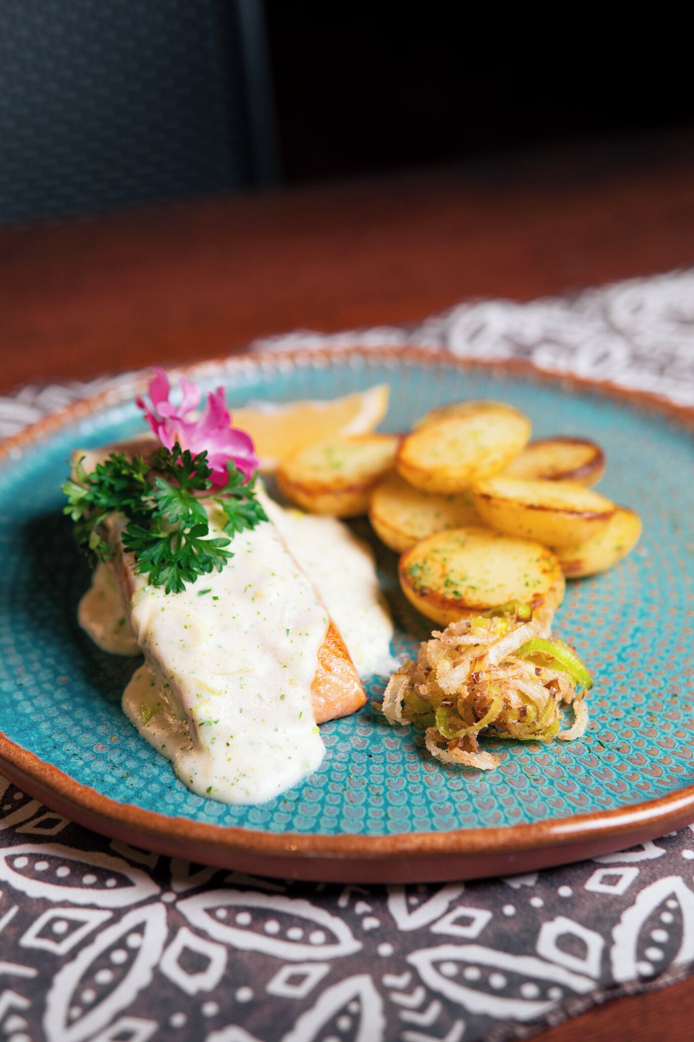 En tallerken med fiskefilet dækket af sauce, pyntet med persille og en lyserød blomst, samt ovnbagte kartoffelskiver.