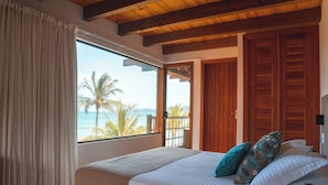 Une chambre avec une grande fenêtre, des poutres en bois au plafond, un lit avec des oreillers et une vue sur les palmiers et la mer.