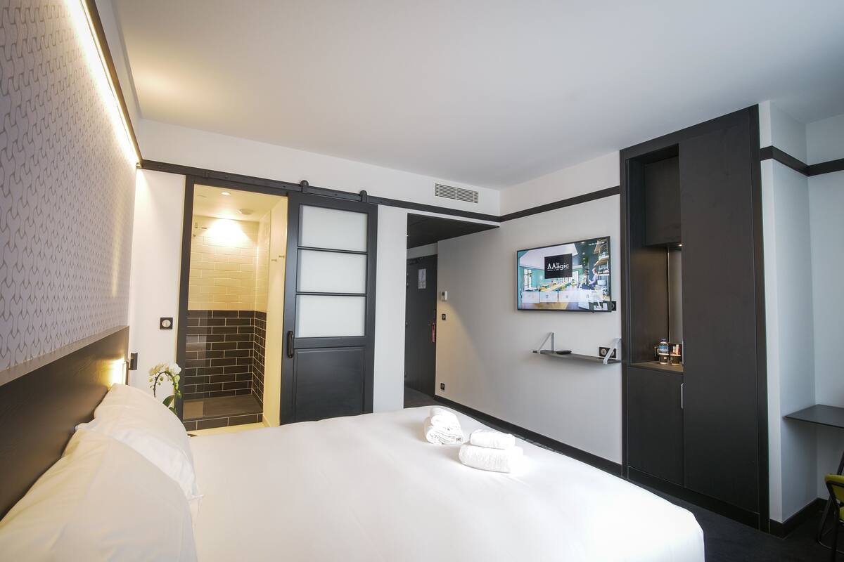 A modern hotel room with a large bed, a TV mounted on the wall, a bathroom with a shower, and a small seating area.