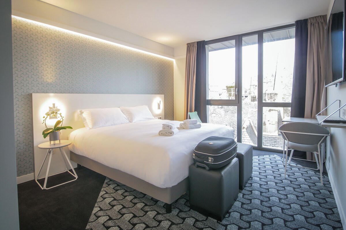 A modern hotel room with a large bed, a desk, and a view of the city.