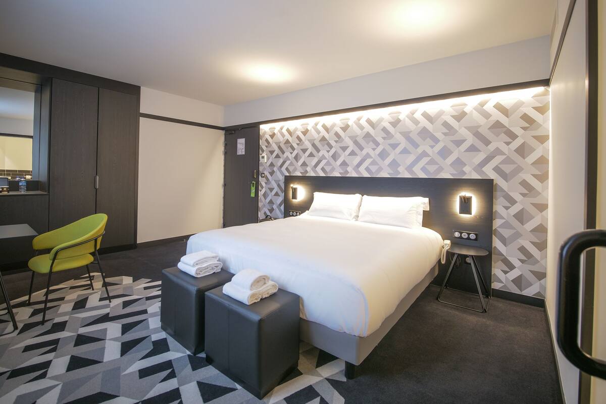 A modern hotel room with a large bed, a desk, a chair, and a patterned carpet.