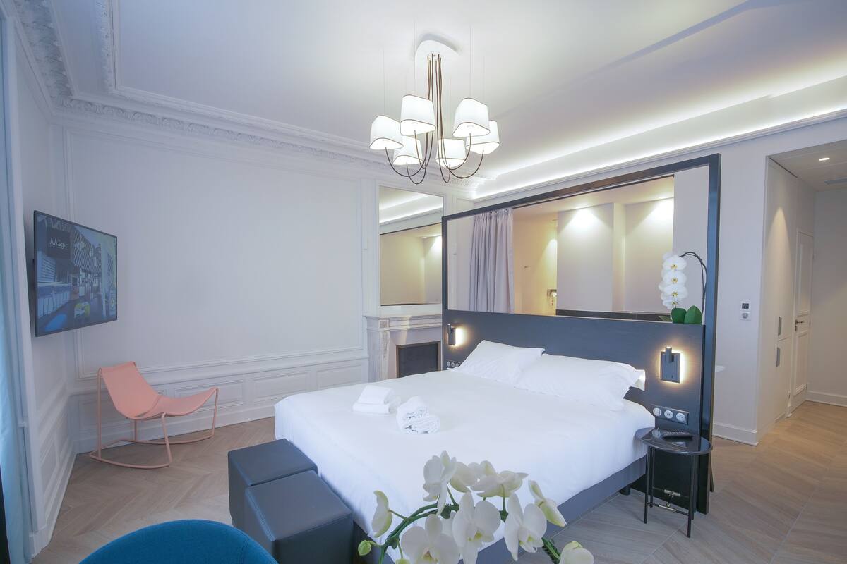 A modern hotel room with a large bed, a sitting area with a chair, a small table, and a mirror.