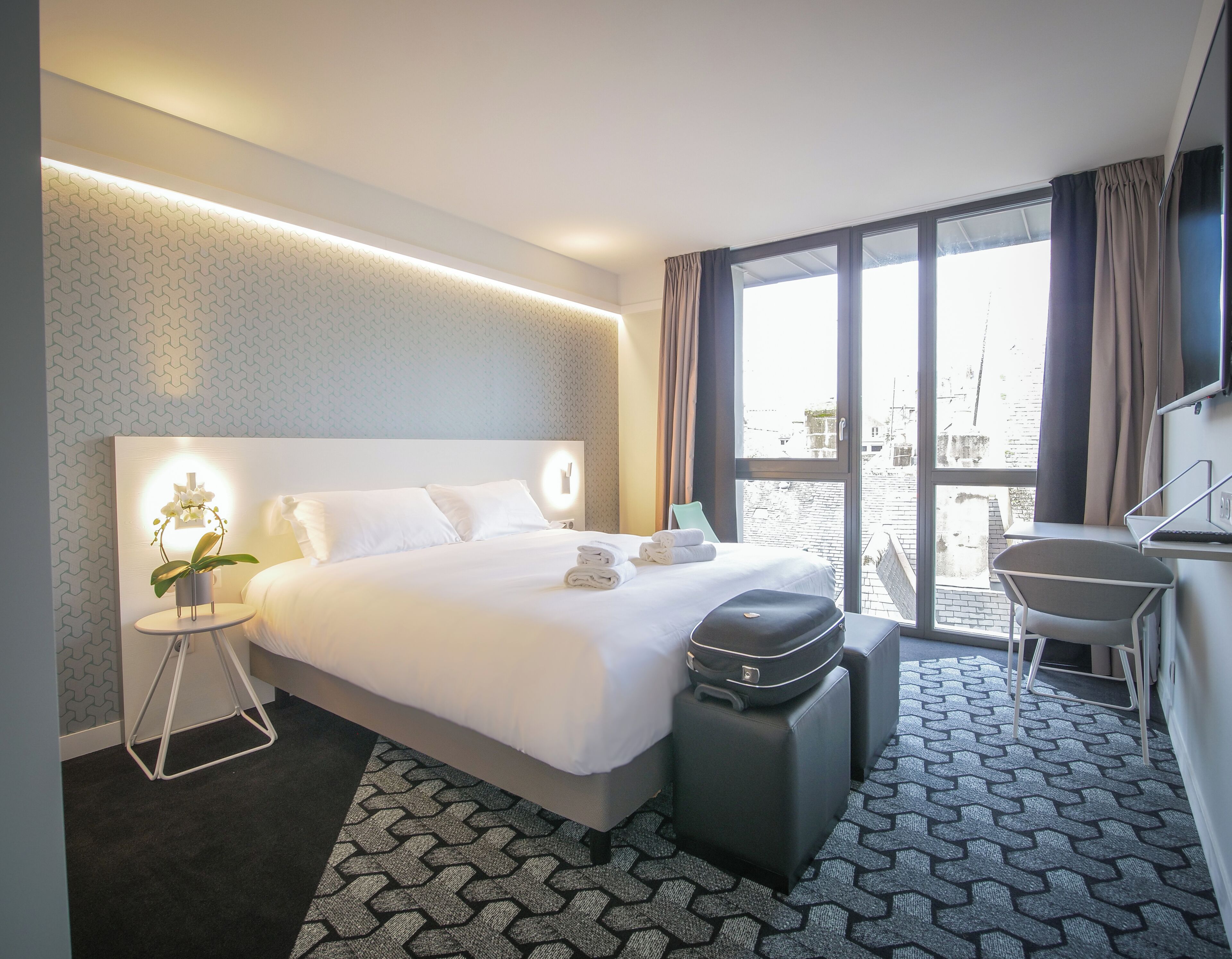 A hotel room with a bed, a desk, a chair, a suitcase, a small table with a plant, and a large window with a view.