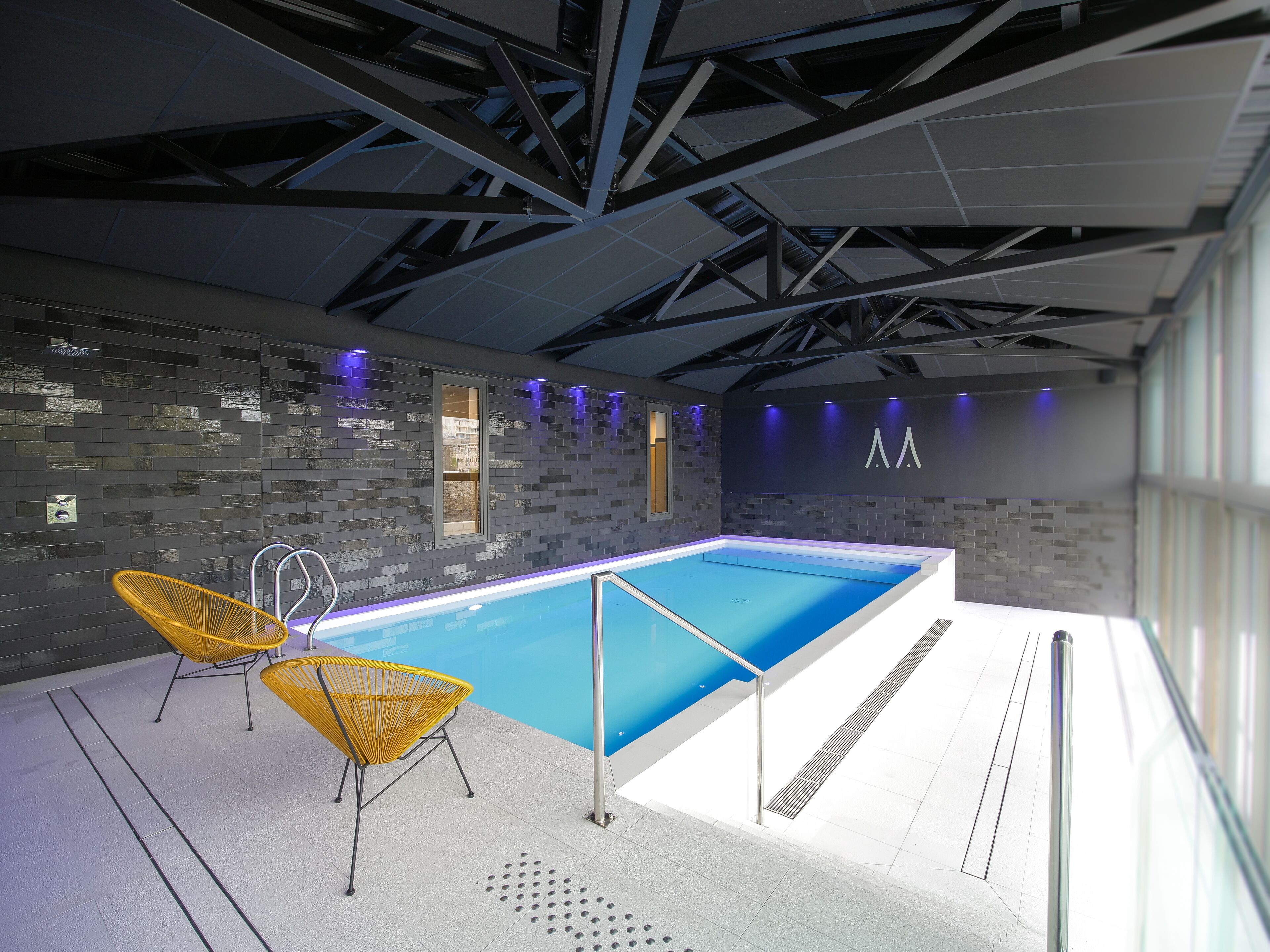 Une piscine intérieure moderne avec un sol carrelé, deux chaises jaunes et un plafond avec des poutres apparentes.