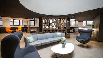 Un espacio lounge moderno con techo curvo, un sofá gris, dos sillones oscuros y una mesa redonda blanca con una planta.