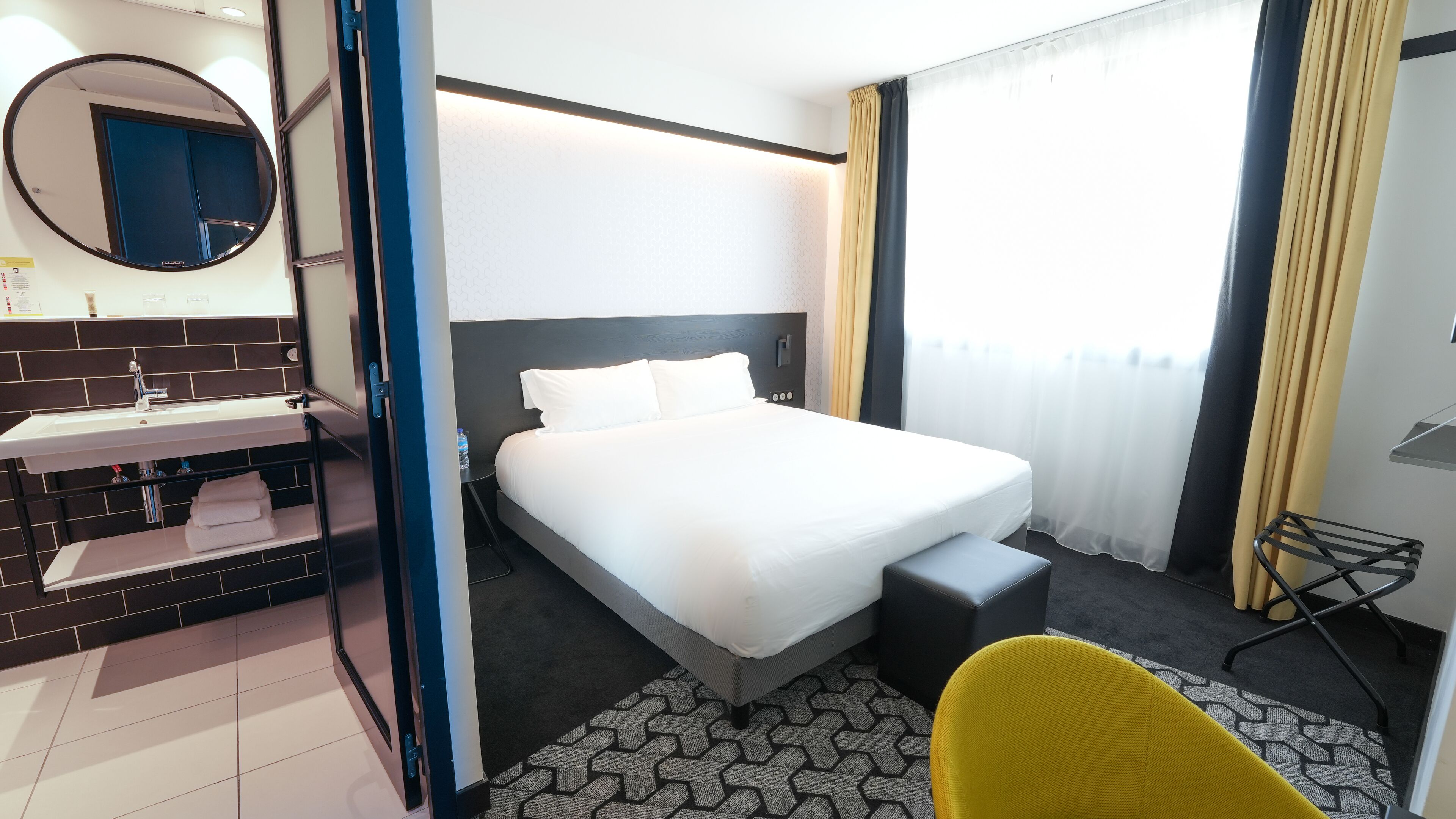 A hotel room with a bed, a bedside table, a chair, a bathroom with a sink, and a mirror.