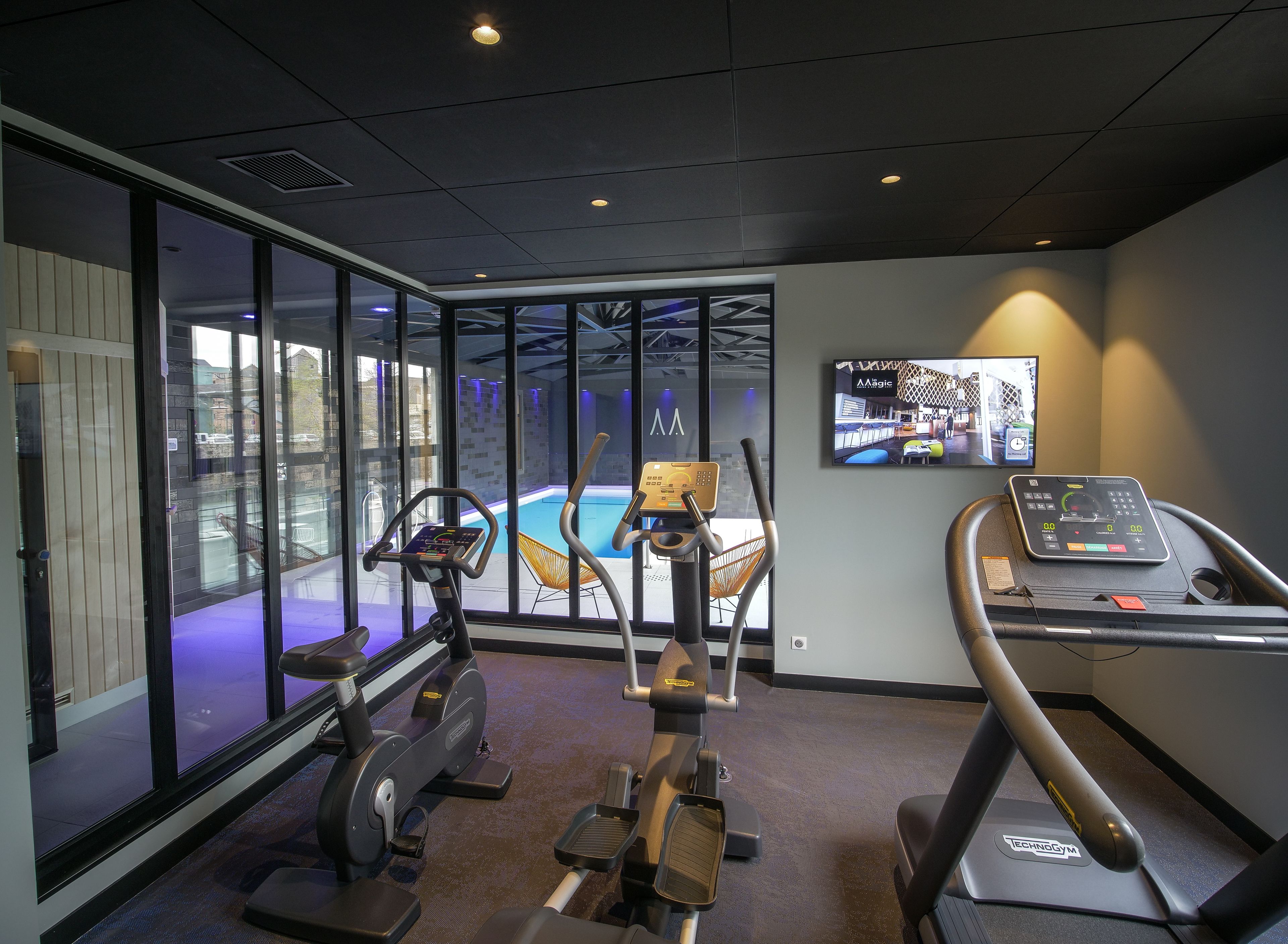 Ein Fitnessraum mit Cardiogeräten, einem Bildschirm und Blick auf einen Pool.