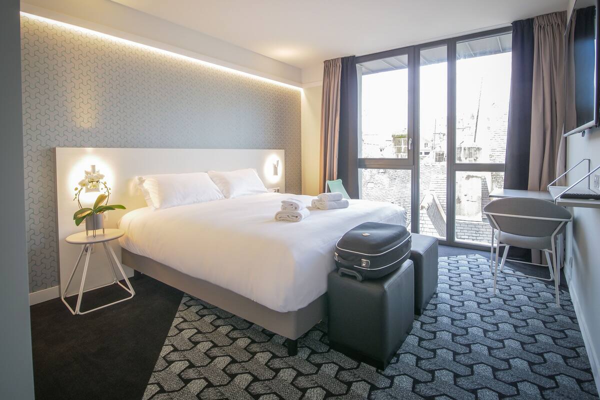 A modern hotel room with a large bed, a desk, and a view of the city.