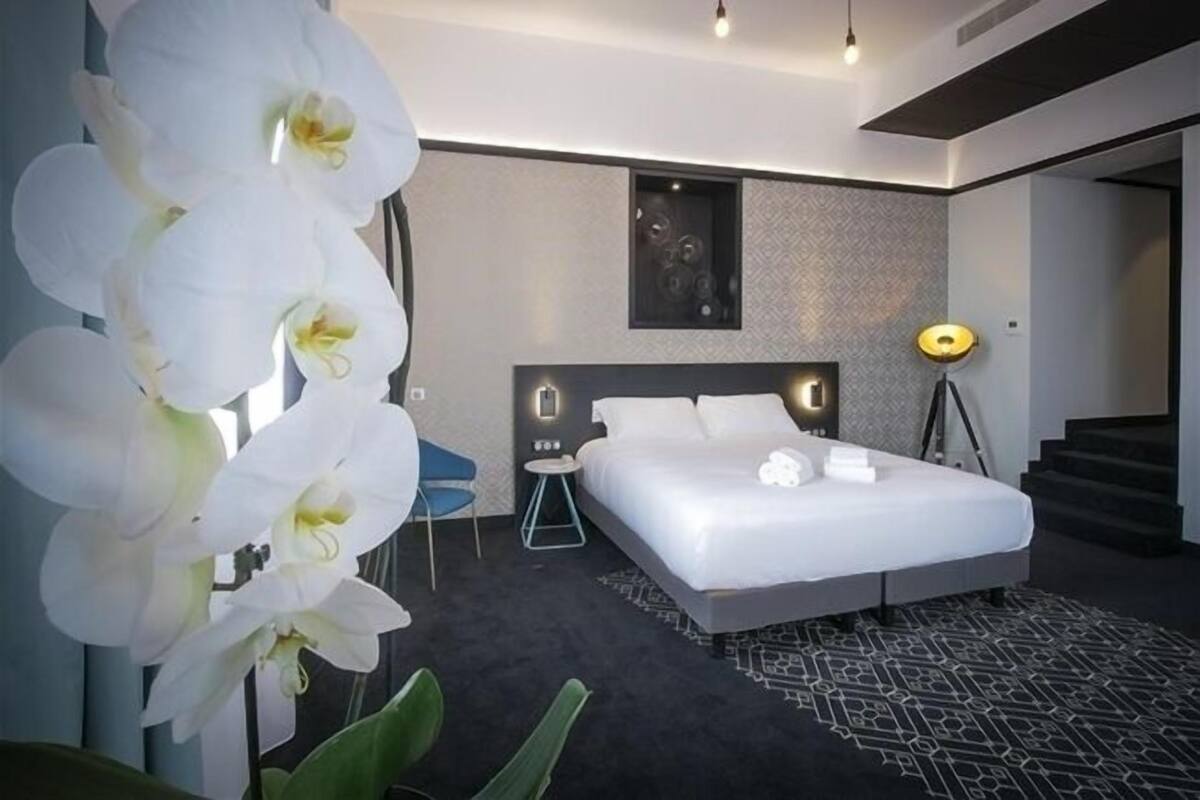 A modern hotel room with a large bed, a desk, and a chair. There is a large window with curtains, a floor lamp, and a plant with white flowers.