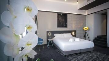 Une chambre d’hôtel moderne, dotée d’un grand lit, d’un bureau et d’une chaise. On y trouve une grande fenêtre avec des rideaux, une lampe de sol et une plante ornée de fleurs blanches.