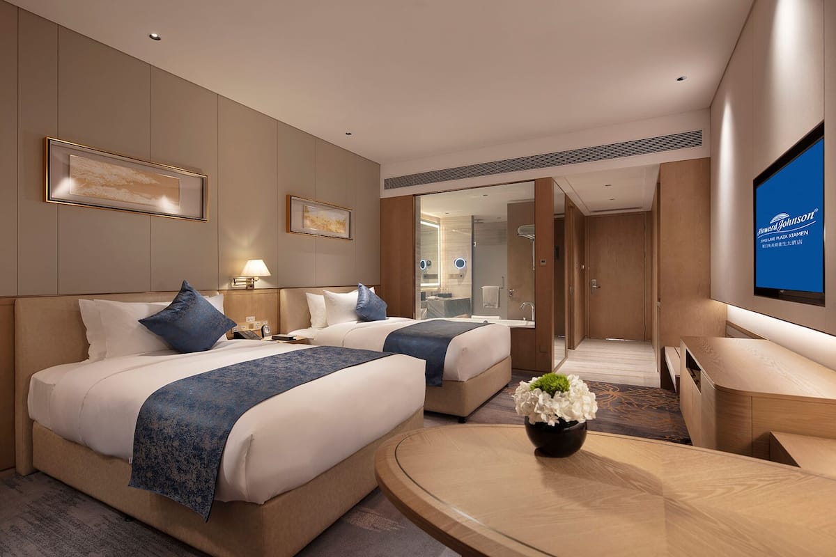 A hotel room with two beds, a desk, a TV, and a bathroom visible through an open door.
