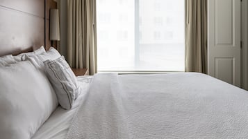 A neatly made bed with white linens and a textured duvet cover, a wooden headboard, and a view of a building through a window with curtains.
