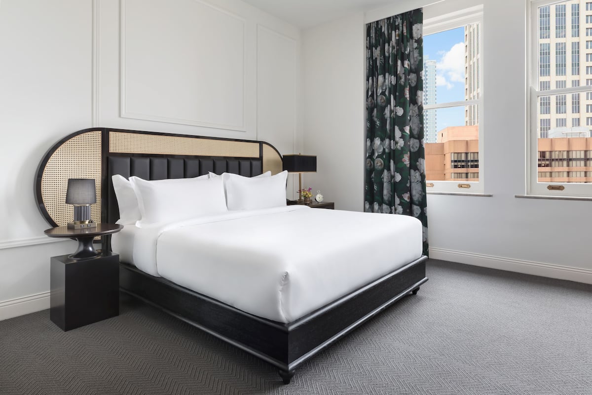 A neatly made bed with a black frame and white bedding, a round side table with a lamp, and a view of buildings through a window with a curtain.
