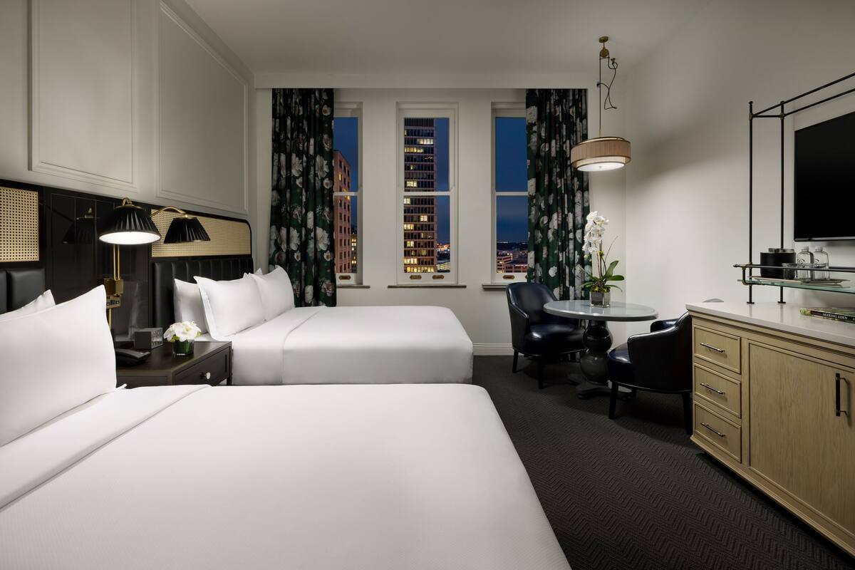 A hotel room with a large bed, a desk with a chair, a TV, and a view of the cityscape through three windows.