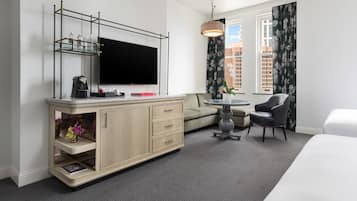 A hotel room with a flat-screen TV, a wooden cabinet with drawers, a round table with a chair, a sofa, and a bed.