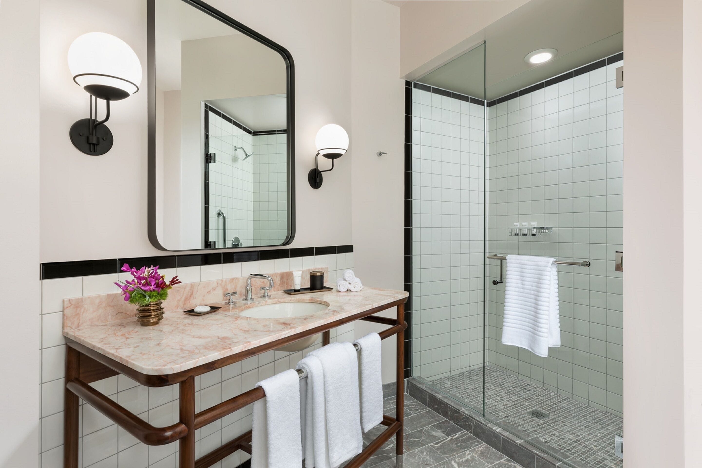 Un baño con lavamanos de mármol, un espejo grande, dos luces empotradas en la pared, una ducha con mampara de vidrio y un perchero para toallas.