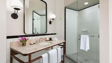 Un baño con lavamanos de mármol, un espejo grande, dos luces empotradas en la pared, una ducha con mampara de vidrio y un perchero para toallas.