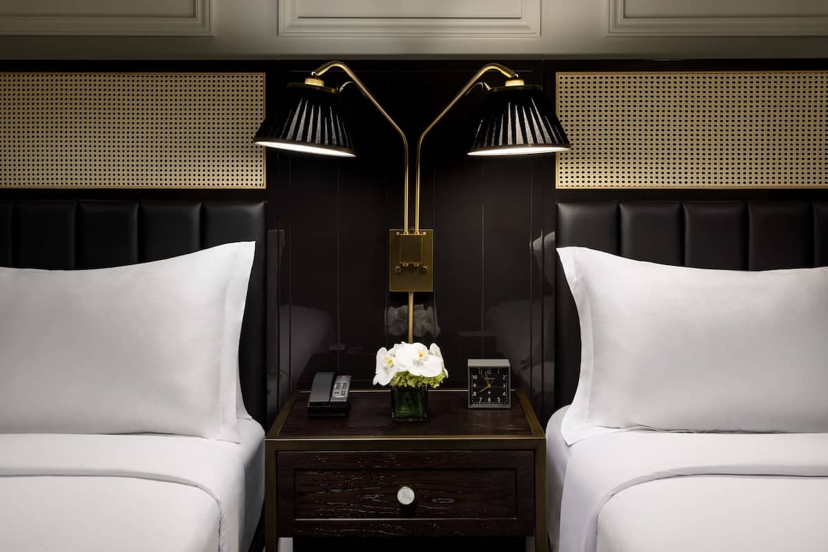 A hotel room with two beds, a nightstand with a lamp, a vase with flowers, a clock, and a phone.