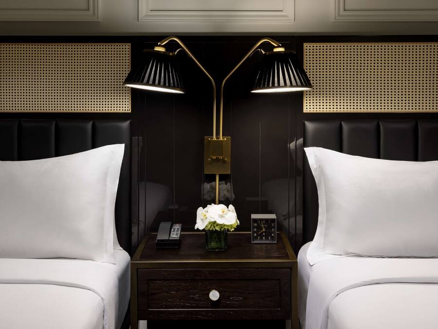 A hotel room with two beds, a nightstand with a lamp, a vase with flowers, a clock, and a phone.