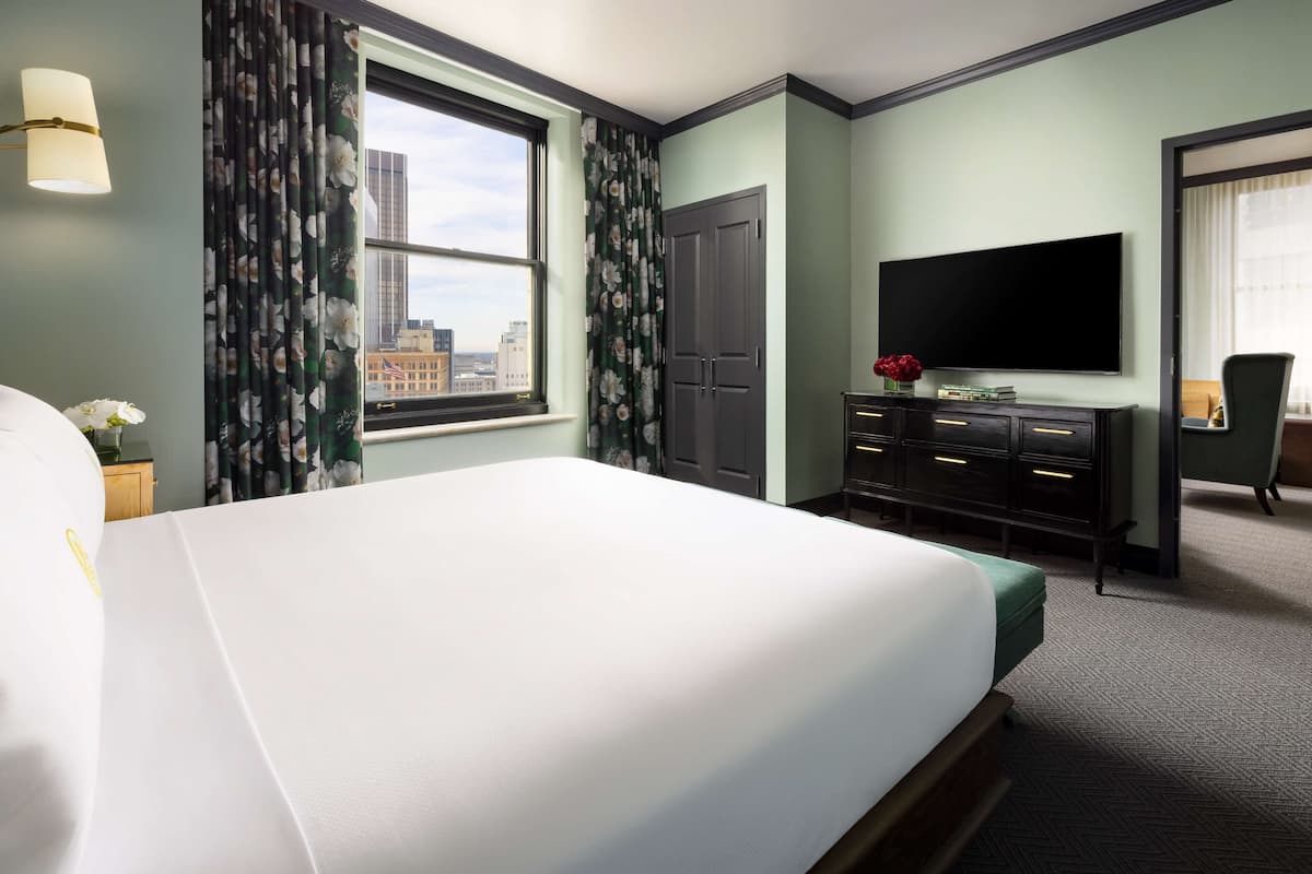 A hotel room with a large bed, a flat-screen TV, a dresser, and a view of the city through a window.