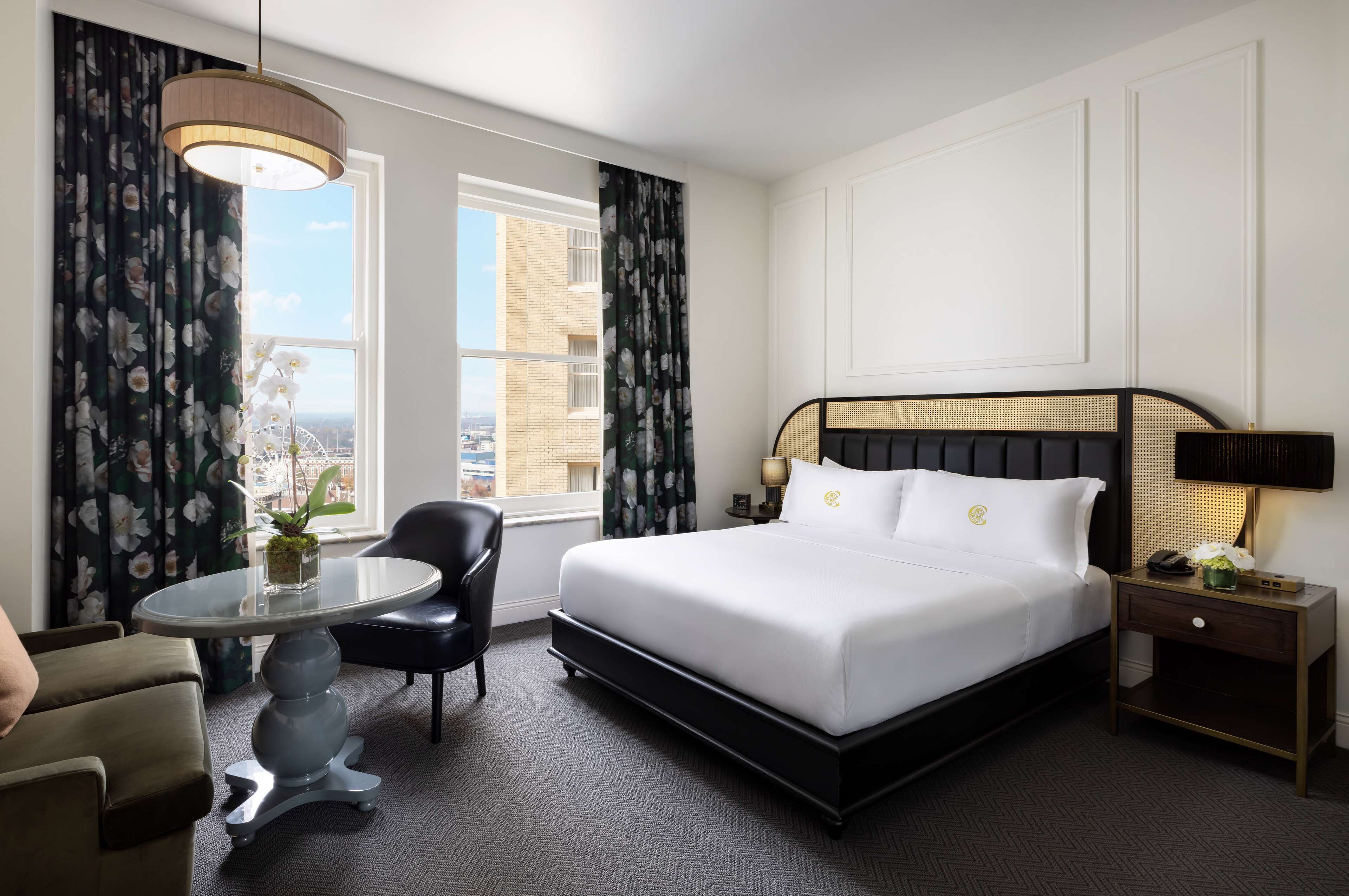 A hotel room with a large bed, a round table, a chair, and a nightstand with a lamp.