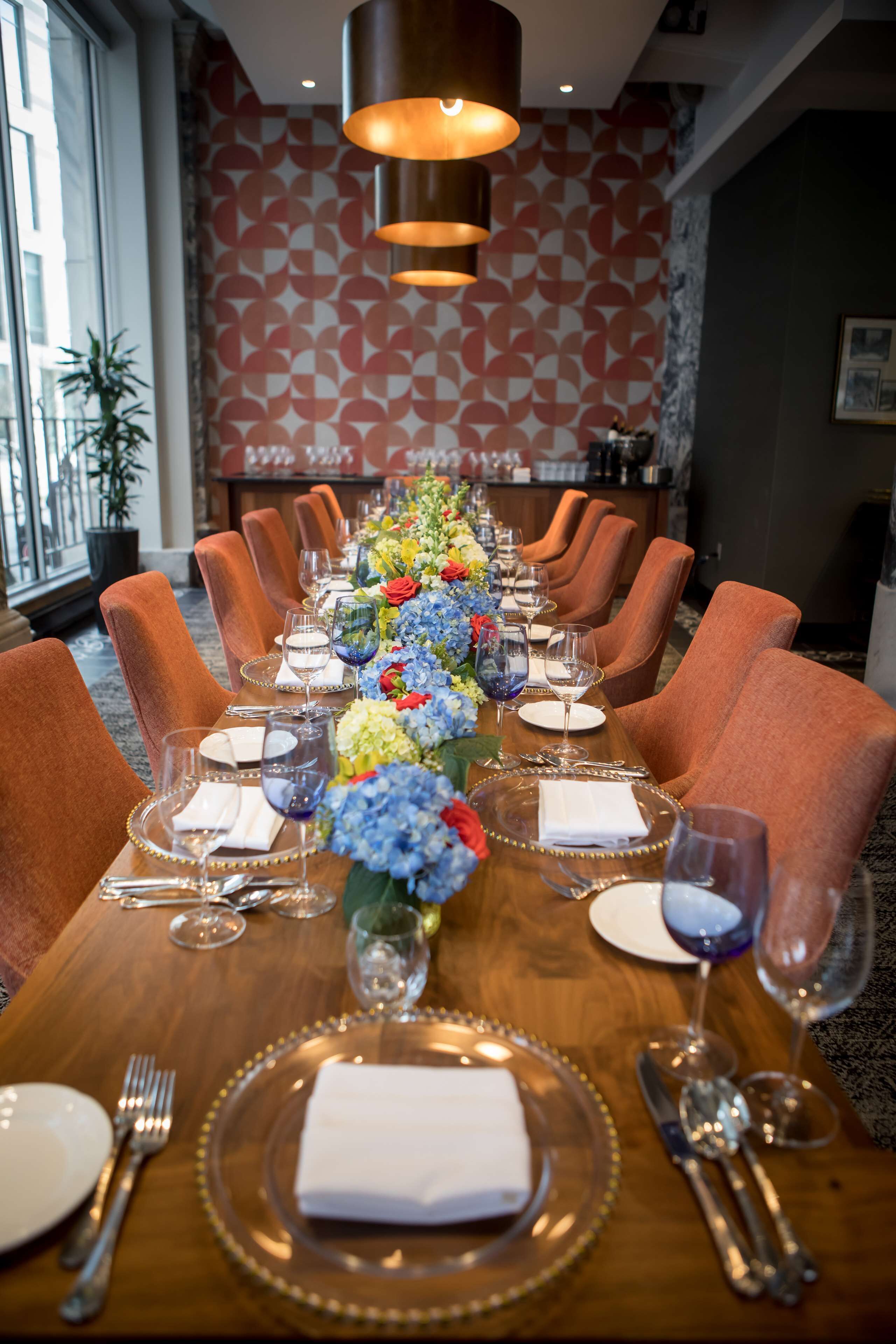 Uma mesa de jantar posta para uma refeição formal, com cadeiras, um arranjo floral no centro e um papel de parede estampado ao fundo.