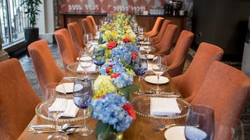 Uma mesa de jantar posta para uma refeição formal, com cadeiras, um arranjo floral no centro e um papel de parede estampado ao fundo.