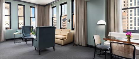 A hotel room with a sofa, armchairs, a small table, a desk, and a chair. There are windows with curtains, a lamp, and a vase with flowers.