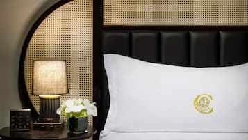 A neatly made bed with a white pillow featuring a logo, a bedside table with a lamp, a clock, and a vase with flowers.