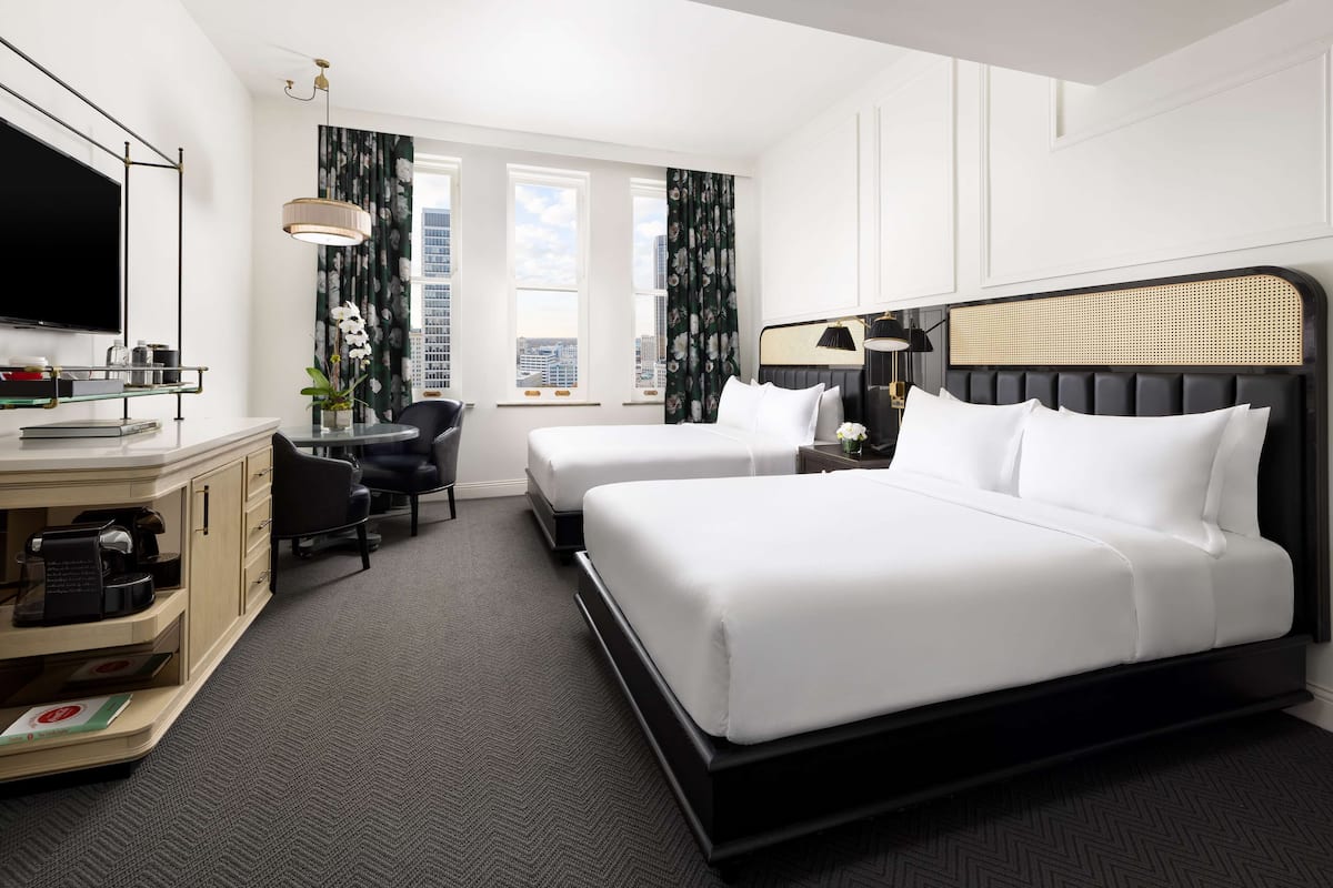 A hotel room with two beds, a TV, a desk with a coffee machine, and a view of the city.