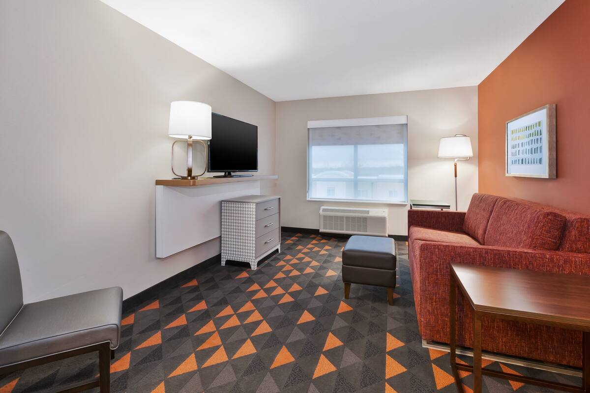 Ein Hotelzimmer mit Fernseher, einer Couch, einem kleinen Tisch, einem Sessel und einem gemusterten Teppich.