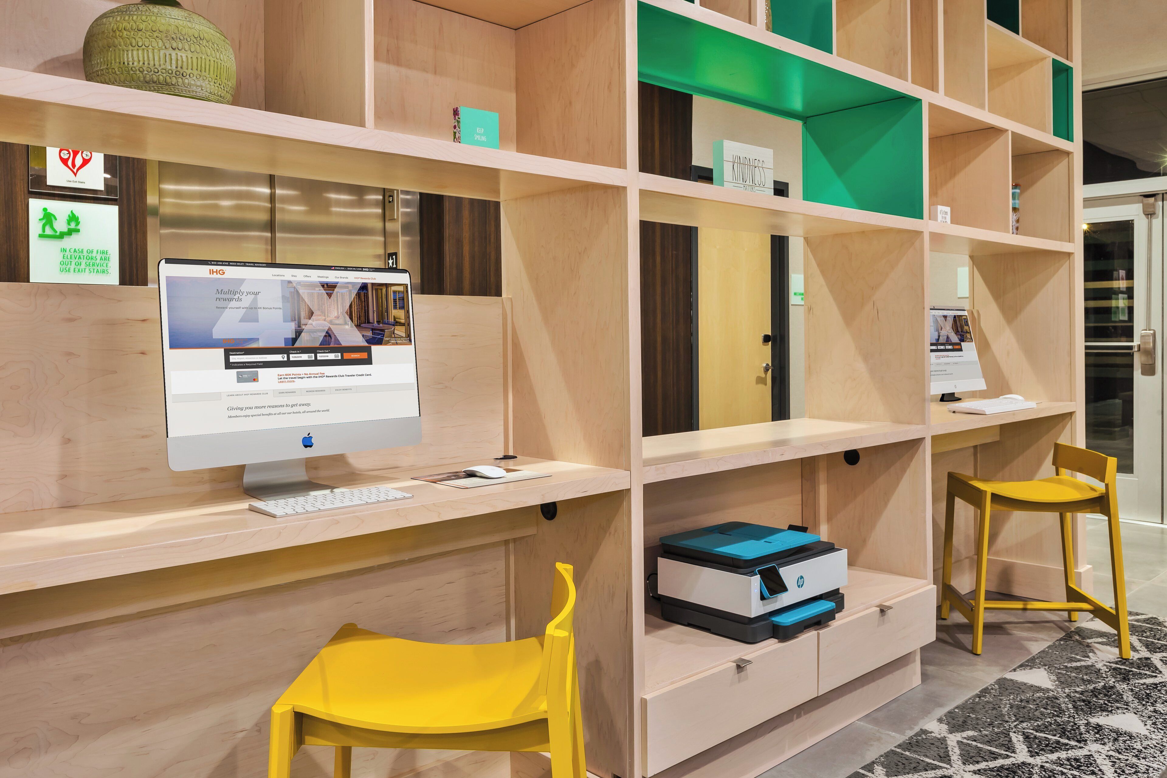 A hotel reception desk with a computer monitor, keyboard, and mouse. There are two yellow chairs and a green vase on the shelf.