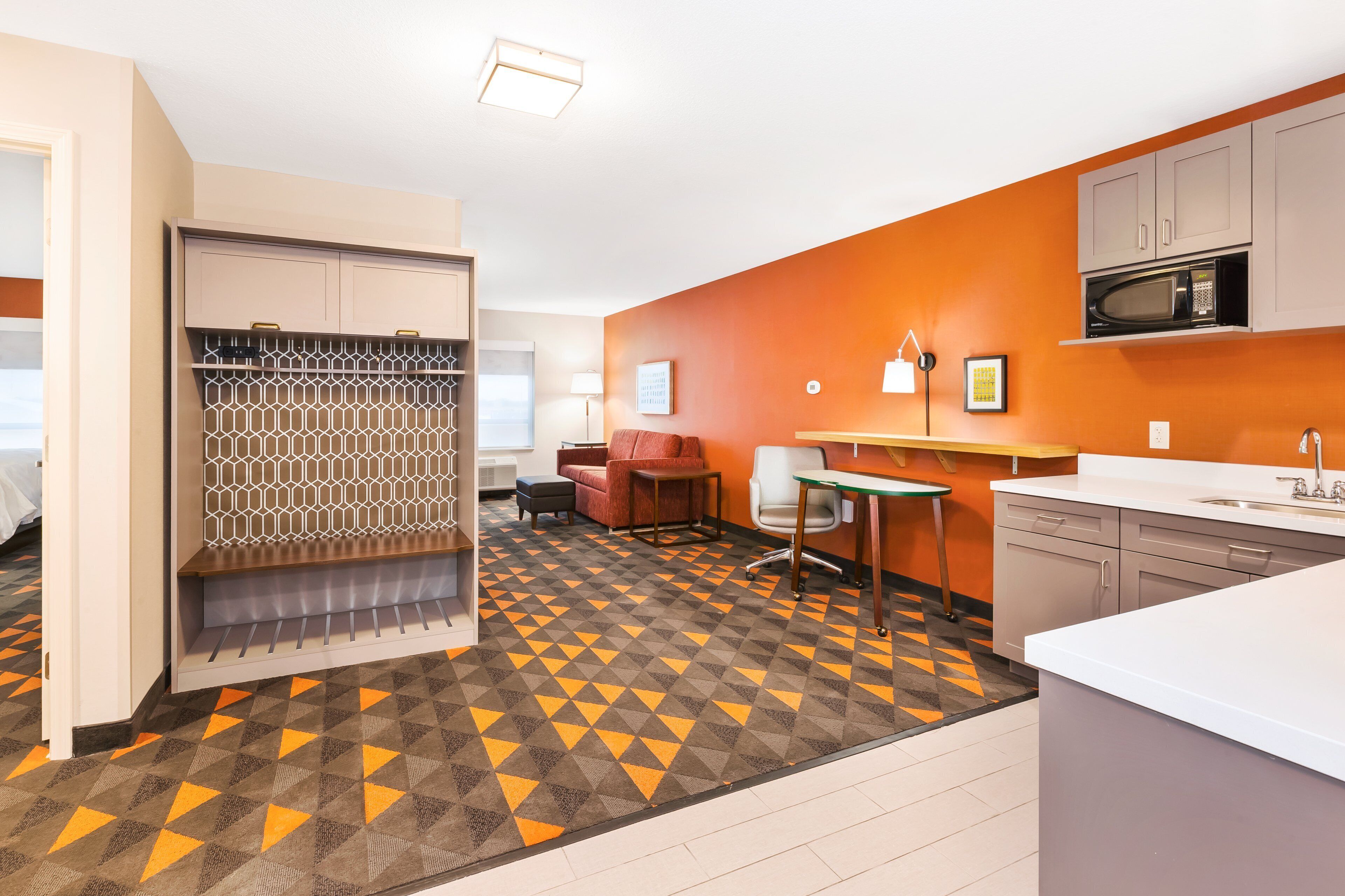 A hotel room with a kitchenette, a seating area with a sofa and armchair, a patterned floor, and a closet with a mirror.