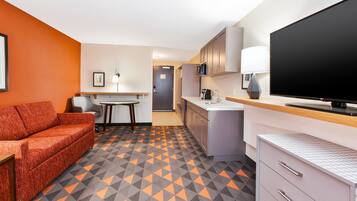 A compact hotel room with a kitchenette, a sofa, a flat-screen TV, and a patterned floor.