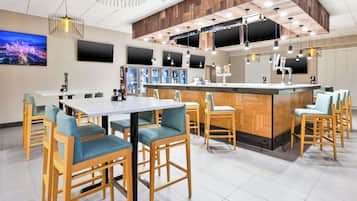 Zona bar moderna con arredi in legno e bancone bar, dotata di schermi multipli e una varietà di bevande.