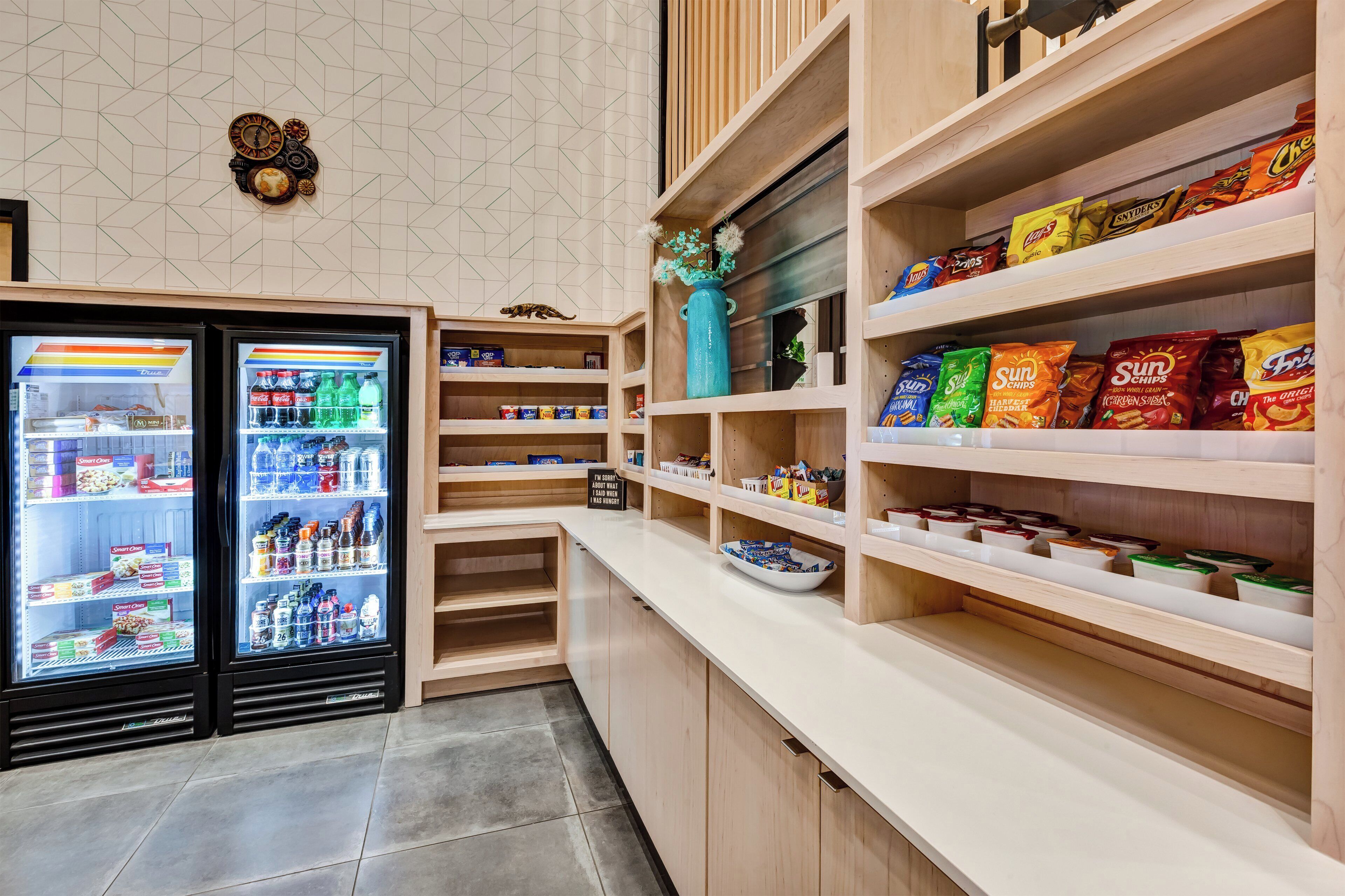 Un bar de aperitivos con estantes llenos de papas fritas y botanas, un refrigerador de bebidas con diversas opciones, y un cartel disculpándose por un error.