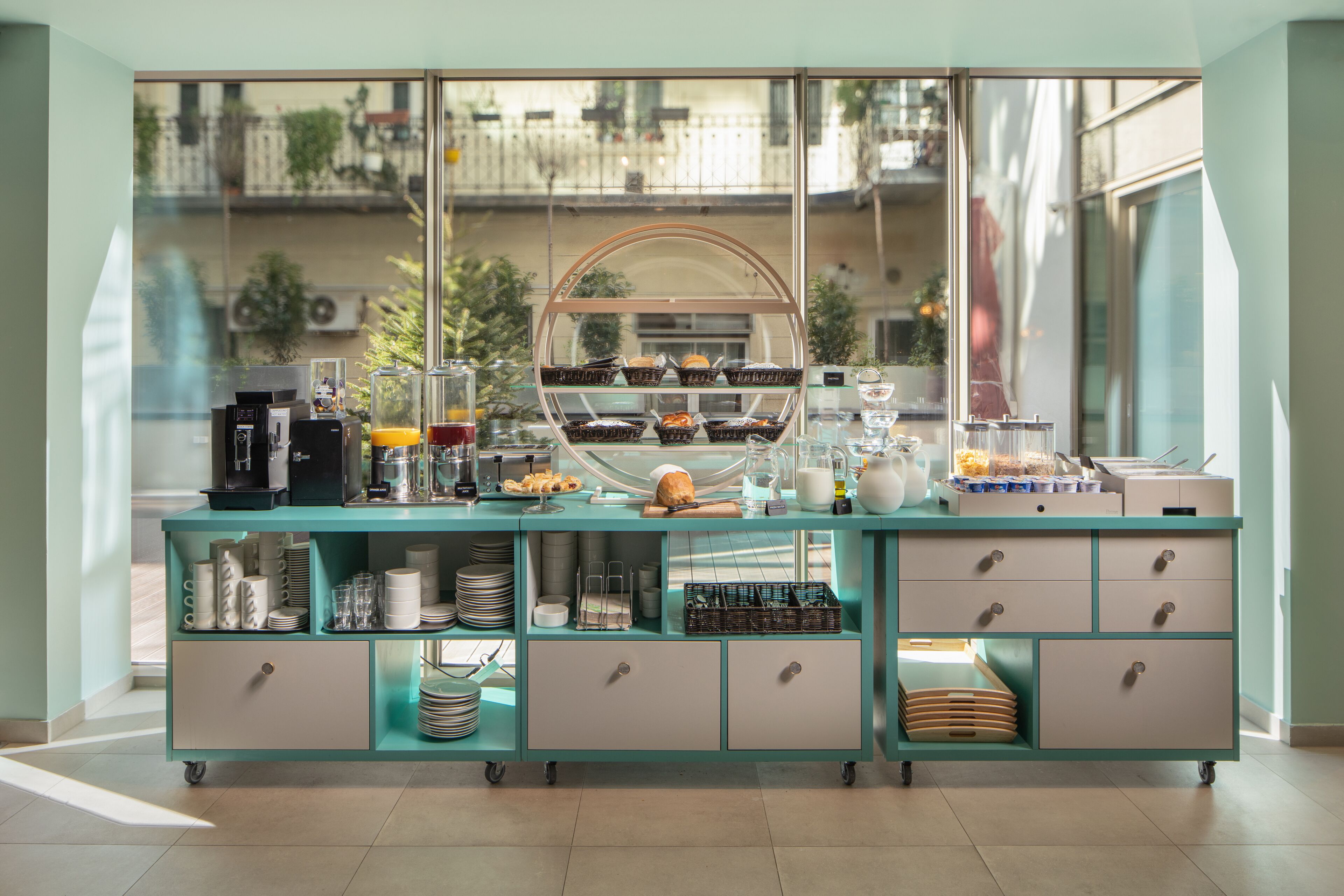 A breakfast buffet with various food items and beverages, a coffee machine, and a view of the outside balcony through large windows.