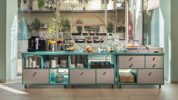 A breakfast buffet with various food items and beverages, a coffee machine, and a view of the outside balcony through large windows.