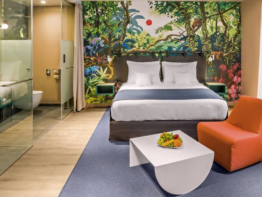 Un dormitorio con cama, una silla, una mesita con fruta y un box de ducha de vidrio, además de un mural con una escena de selva.