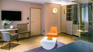 A modern hotel room with a flat-screen TV, a desk with a chair, a sofa, a glass-enclosed shower area, and a small fruit bowl on the coffee table.