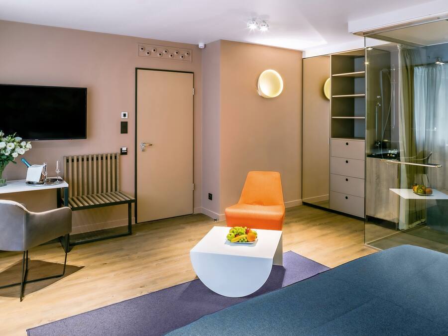 A modern hotel room with a flat-screen TV, a desk with a chair, a sofa, a glass-enclosed shower area, and a small fruit bowl on the coffee table.