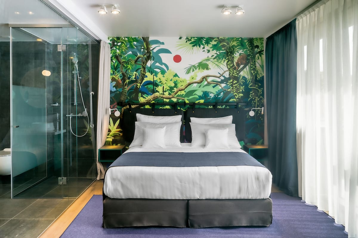 A modern hotel room with a large bed, a glass shower enclosure, and a tropical mural on the wall.