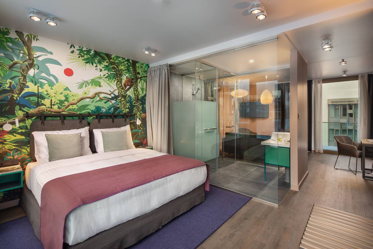A modern hotel room with a large bed, a glass shower enclosure, a dining area, and a mural of a jungle scene on the wall.