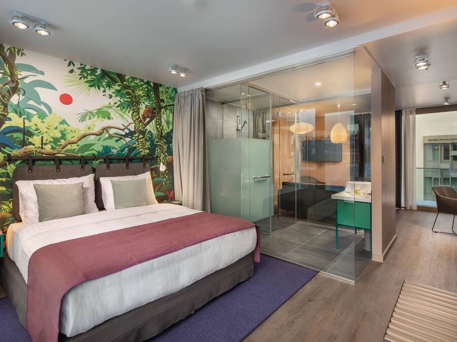 A modern hotel room with a large bed, a glass shower enclosure, a dining area, and a mural of a jungle scene on the wall.