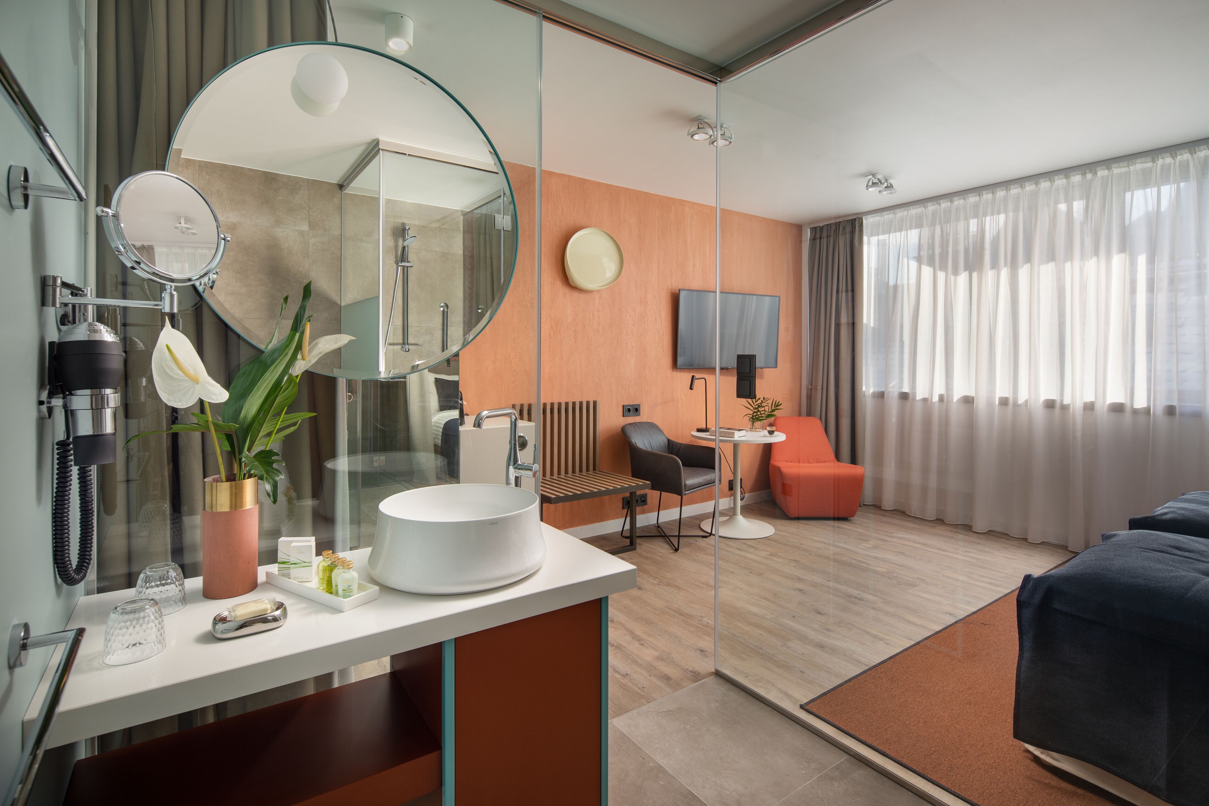 Et moderne hotelværelse med et stort rundt spejl, en hvid vask, et rundt bord med en stol, en seng og et fjernsyn på væggen.