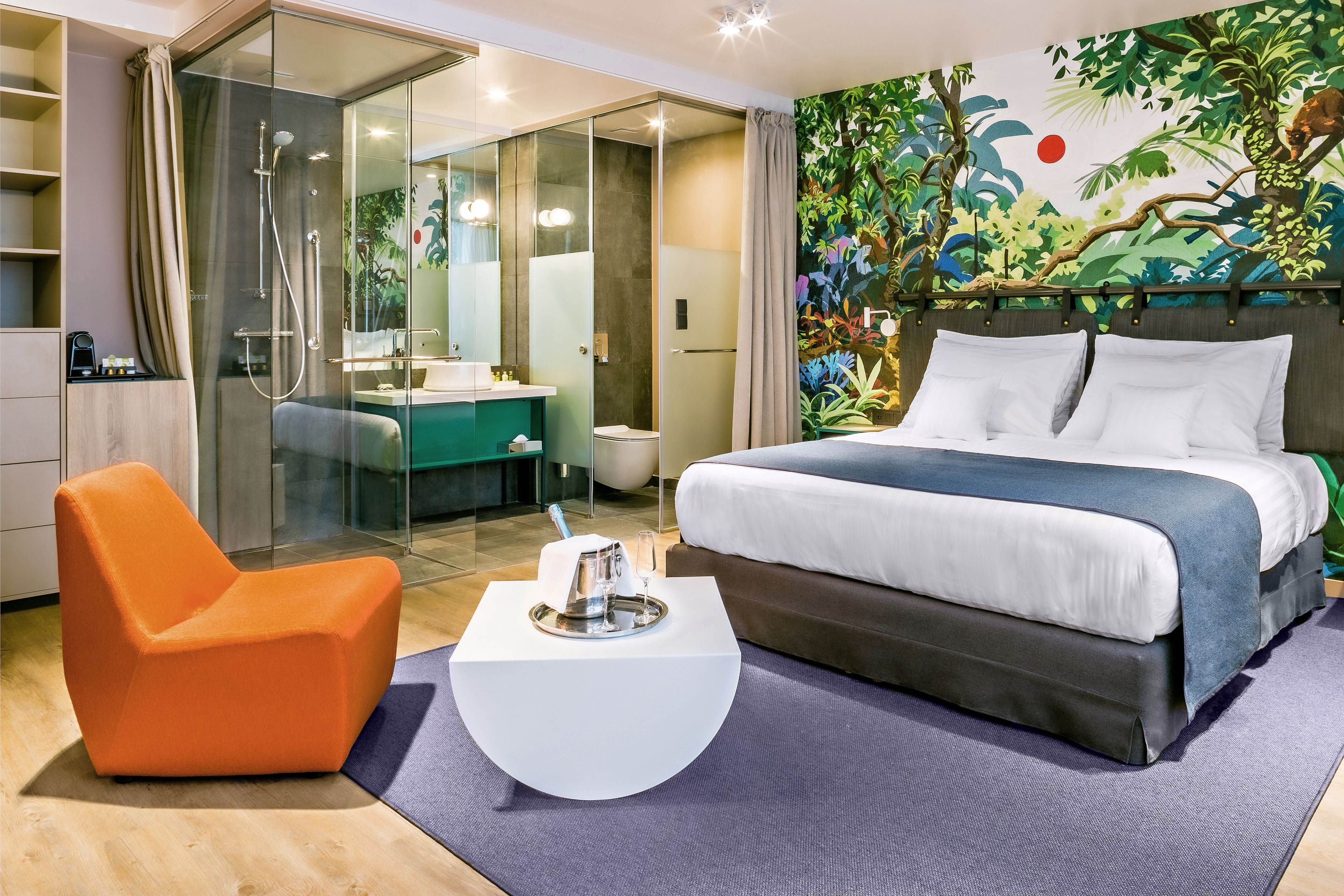 A modern hotel room with a large bed, a glass-enclosed shower, a bright bathroom, and a vibrant jungle-themed wall mural.