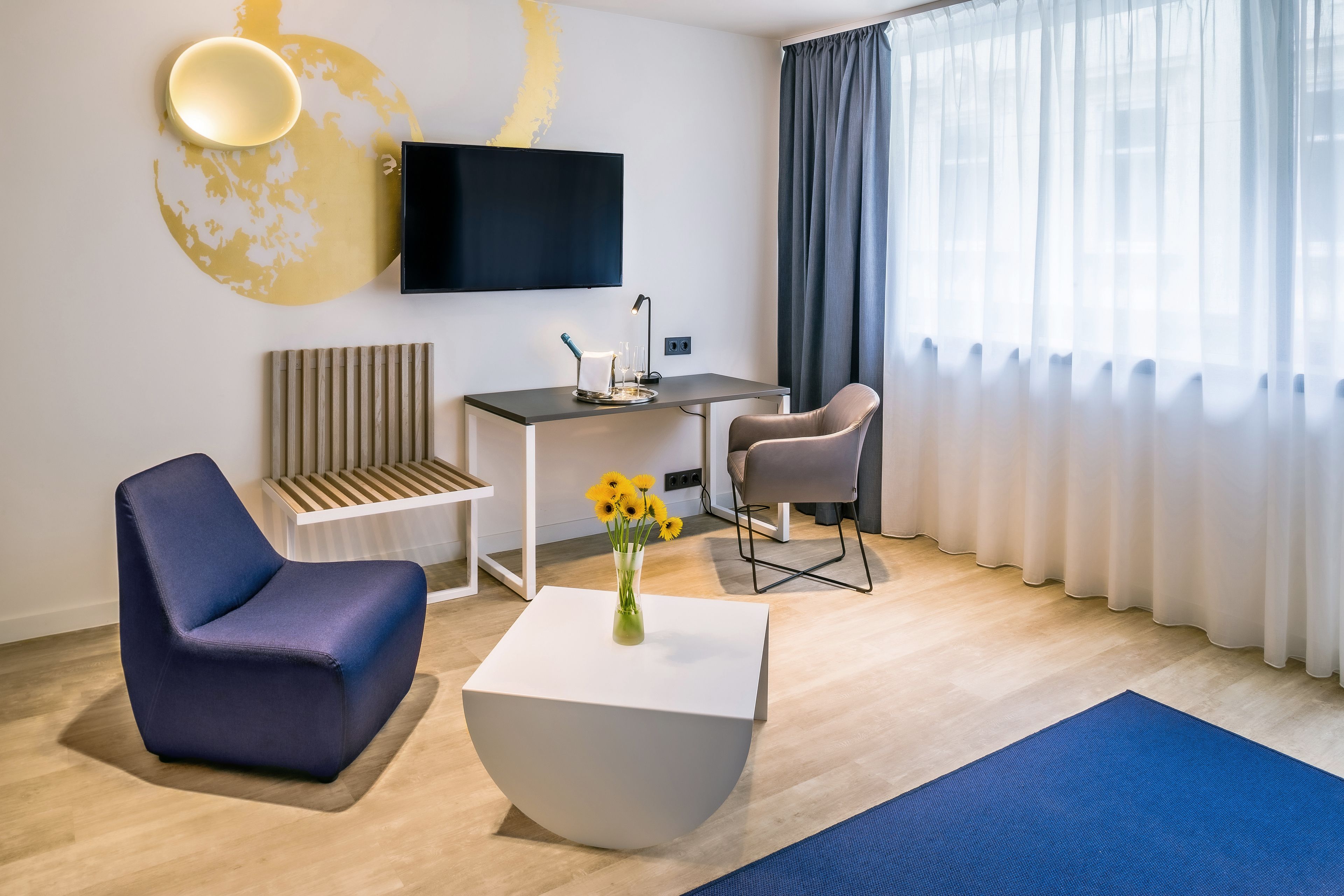 A modern hotel room with a flat-screen TV, a desk, two chairs, a white coffee table, and a large window with sheer curtains.