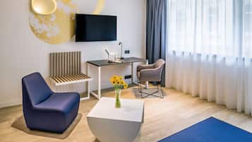 A modern hotel room with a flat-screen TV, a desk, two chairs, a white coffee table, and a large window with sheer curtains.