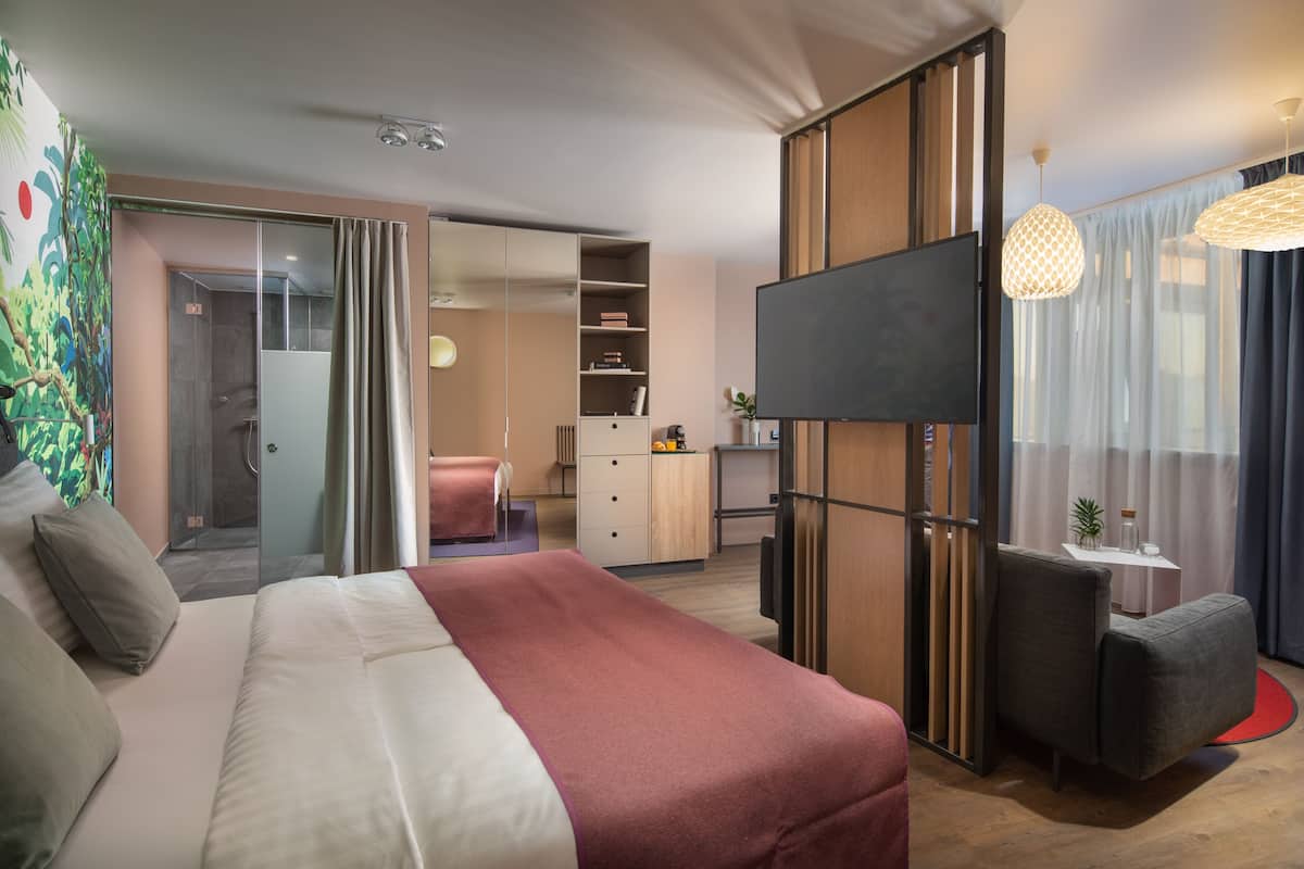 A modern hotel room with a large bed, a TV, a desk, and a sliding door leading to a shower area.